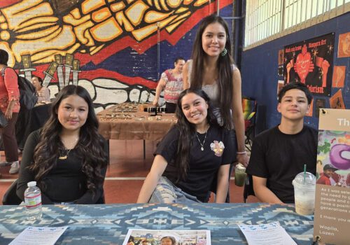 Two young women and one young man sitting at a table with another' young woman standing behind them all smiling