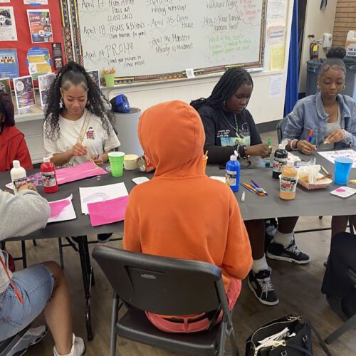 a group of young men and women sitting at a crafting table cutting, cluing things to paper